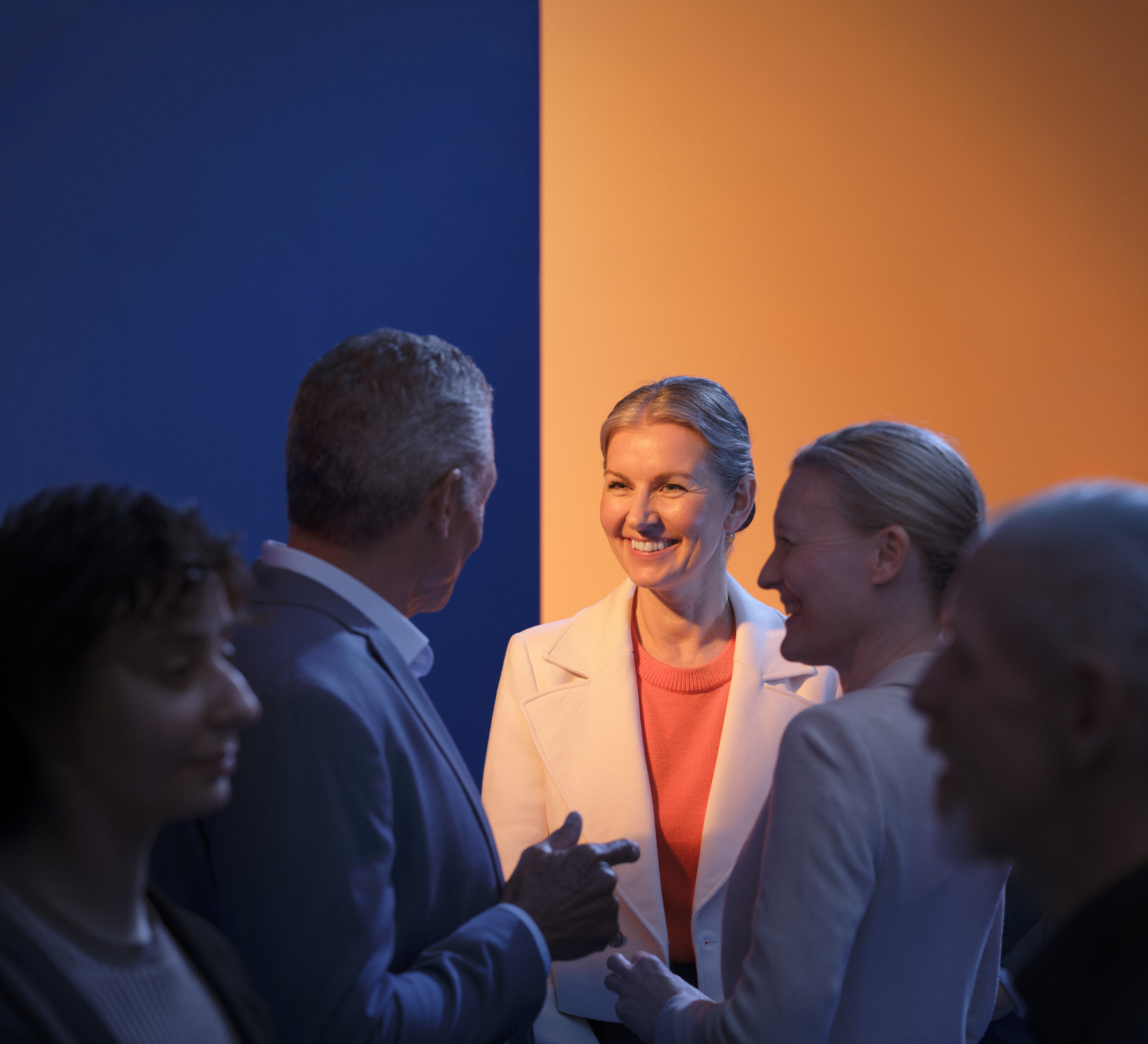Group of people in conversation at an event, with one person in a white blazer and bright orange top standing between a blue and orange background