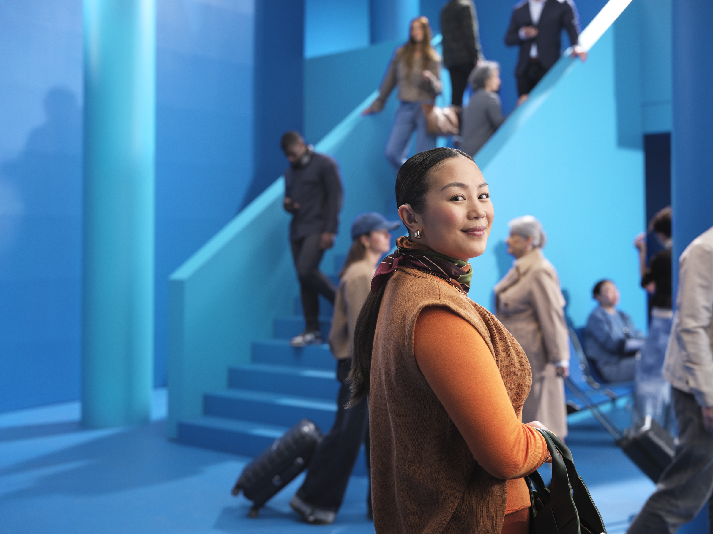 A young woman wearing Beltone Boost Max S hearing aids at a busy station. She is looking comfortably into the camera without being distracted by surrounding sounds using the Clear Focus feature.